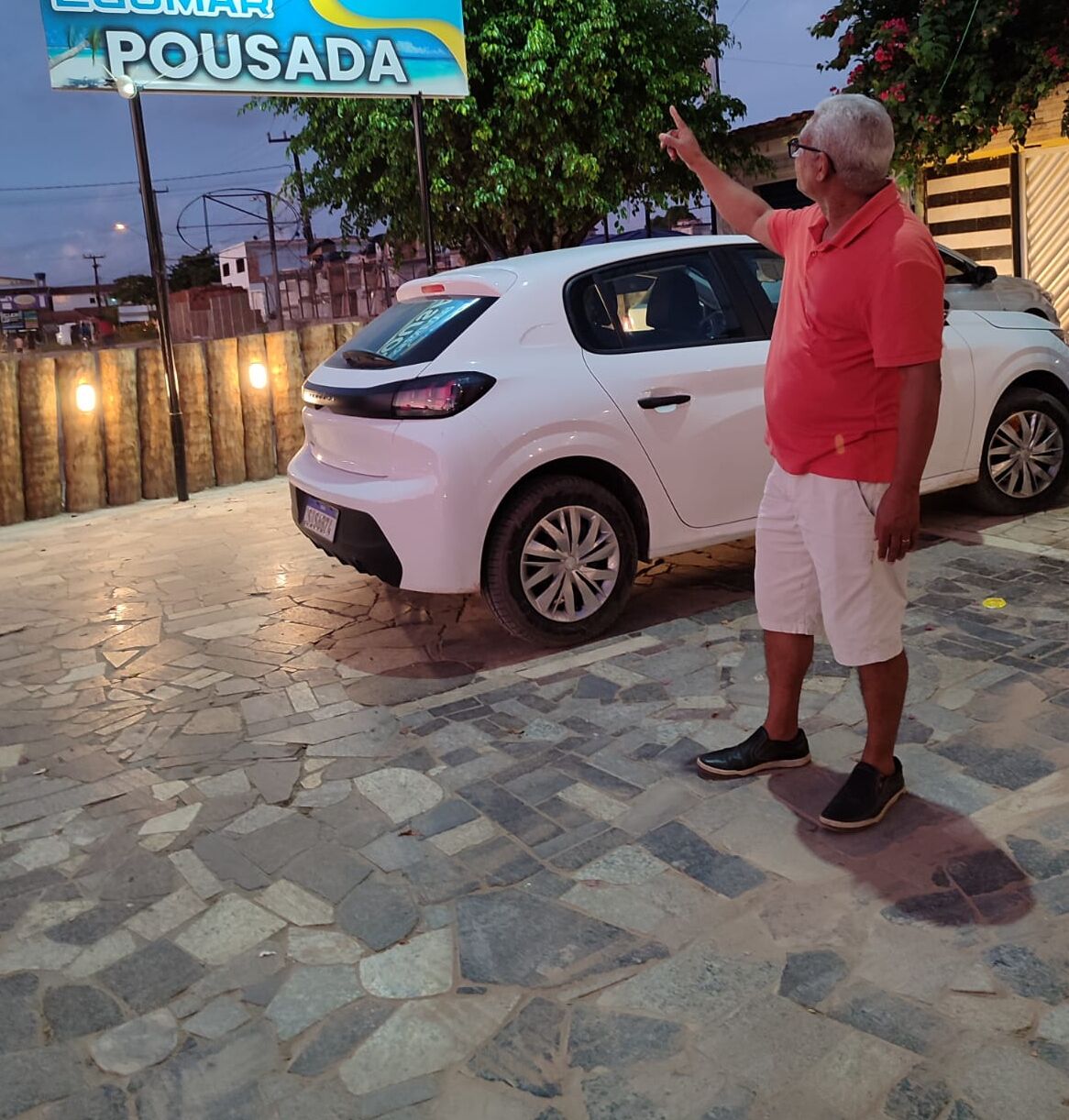 A man standing next to a white car in front of a sign that reads "POUSADA".