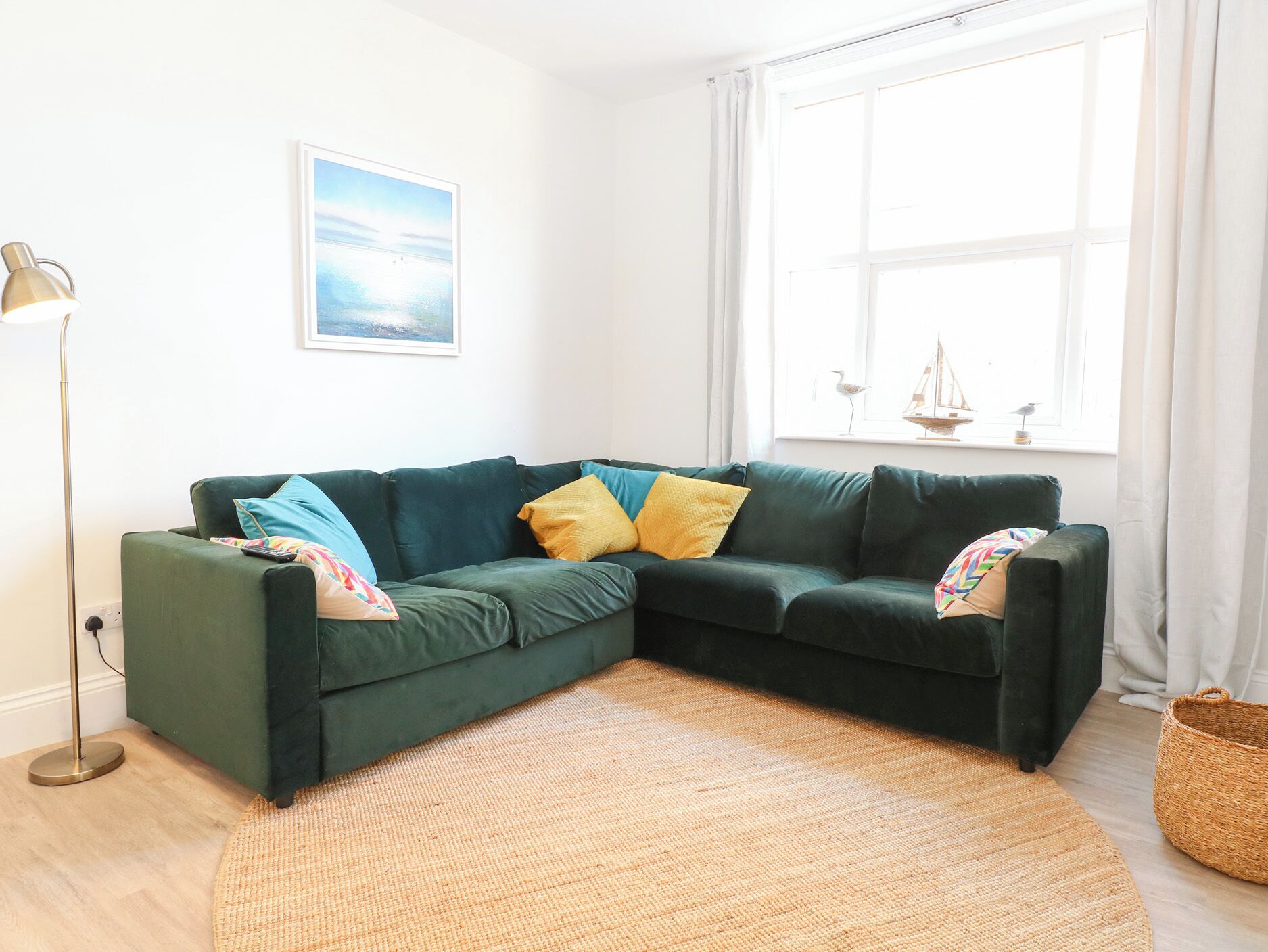 A green sectional sofa with yellow and blue cushions, a woven rug, a floor lamp, and a window with curtains.