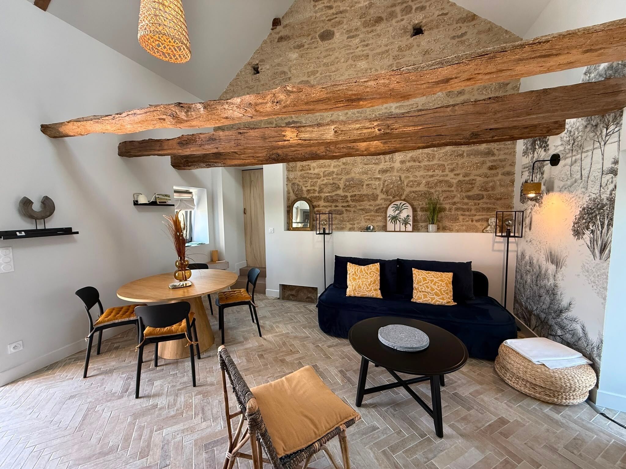 A rustic interior with exposed wooden beams, a stone wall, a round dining table, and a dark blue sofa with patterned cushions.