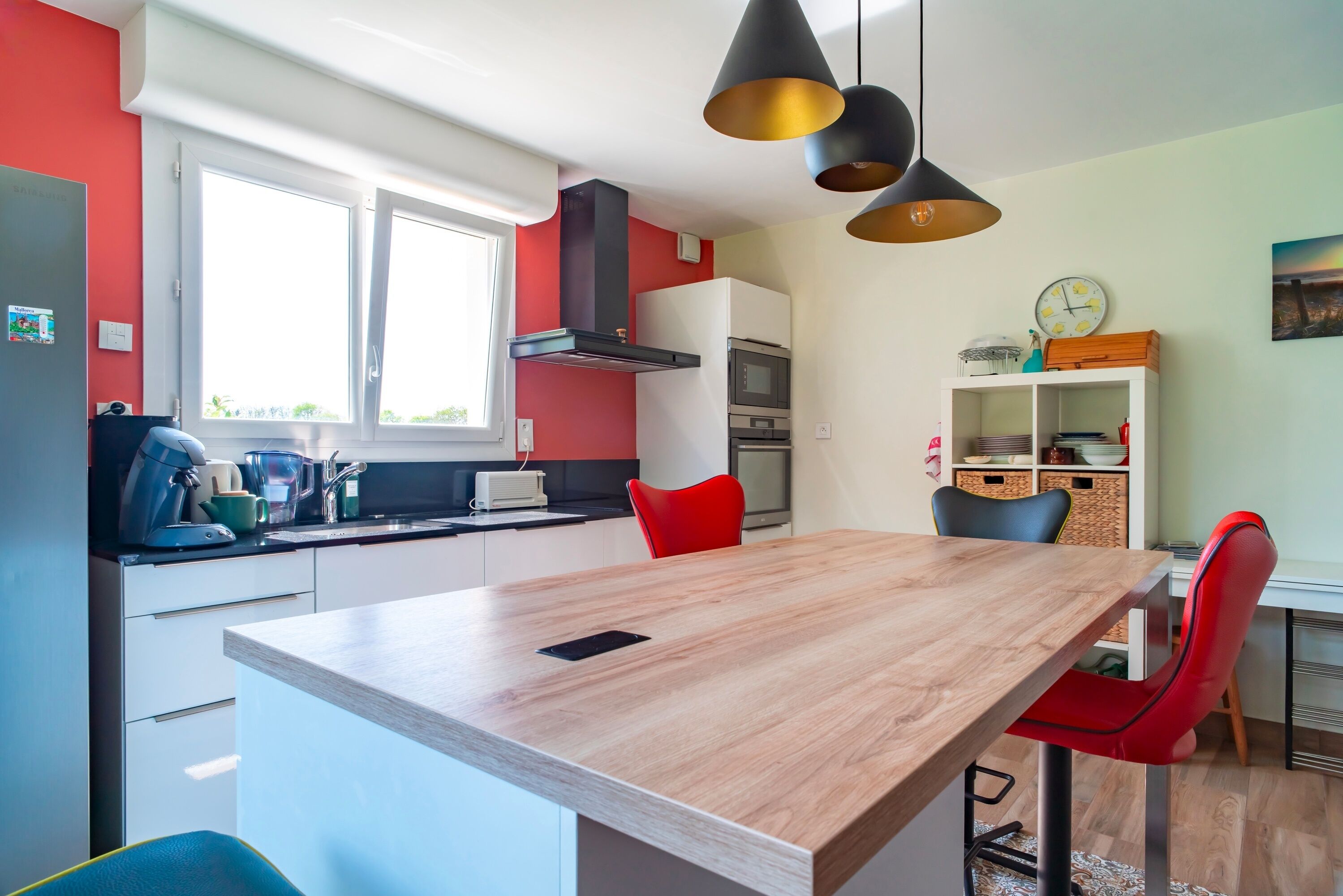 A modern kitchen with white cabinets, a wooden island, red chairs, and black pendant lights.