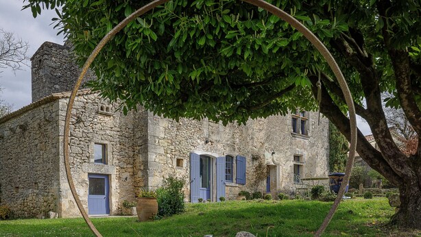 A stone building with a blue door and shutters, surrounded by greenery and a tree.