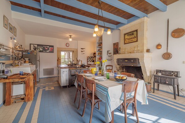 Une cuisine rustique avec des poutres en bois, un coin repas avec une table dressée pour quatre personnes et un poêle d’époque.