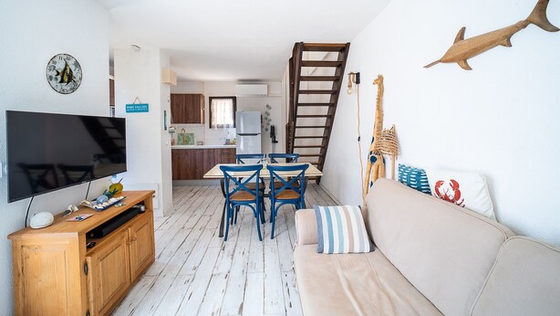 A living room with a TV, wooden entertainment unit, blue chairs, a beige sofa, and a kitchen in the background. - L’Échappée Méditerranéenne - Proche Plage (Leucate)