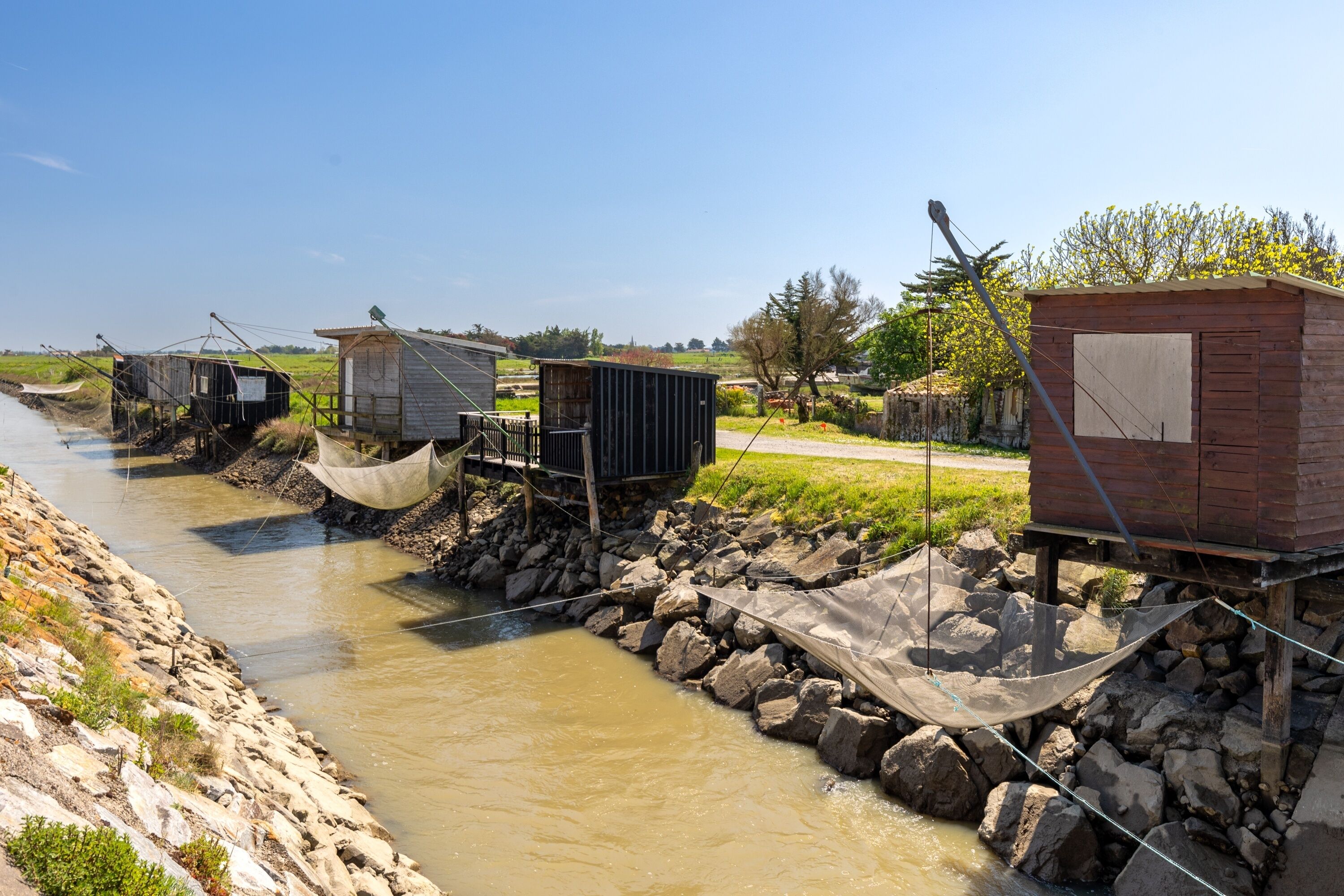 Une rive de rivière bordée de plusieurs constructions en bois, dont certaines sont surélevées sur des piliers, et qui servaient probablement à la pêche ou au stockage.