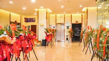 A hotel lobby with a reception desk, a glass partition, and multiple flower arrangements on stands.