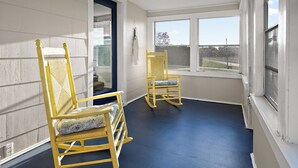 A room with two rocking chairs, a door with a 'Welcome' sign, and a view through large windows.