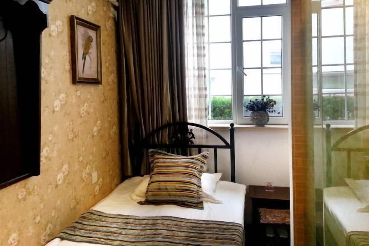 Ein Schlafzimmer mit Bett, einem Fenster mit Vorhängen, einem gerahmten Bild an der Wand und einem kleinen Tisch mit einer Pflanze.