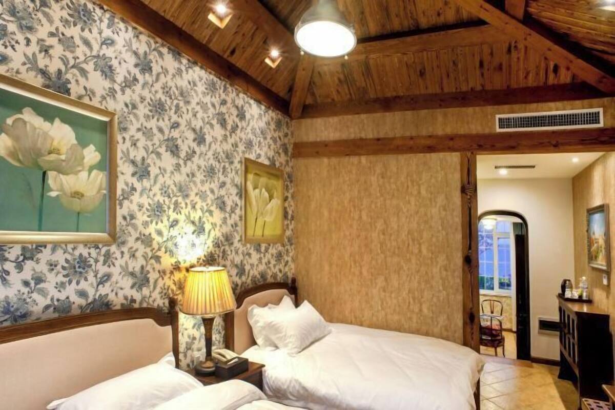 Ein Hotelzimmer mit Holzbalkendecke, Blumenwallpaper, einem Bett mit weißen Leinentüchern, einer Nachttischlampe und einem gerahmten Bild an der Wand.