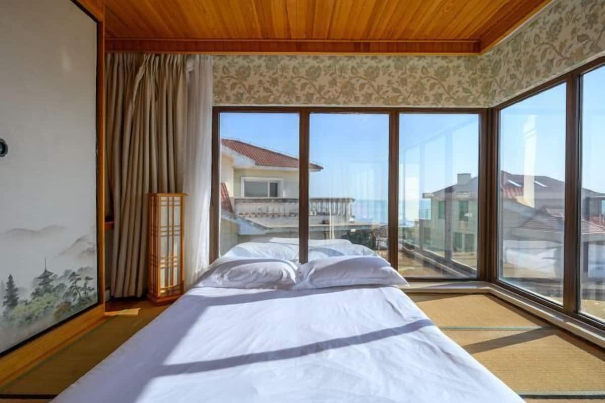 Ein Zimmer mit einem Bett, großen Fenstern und Blick auf Gebäude.