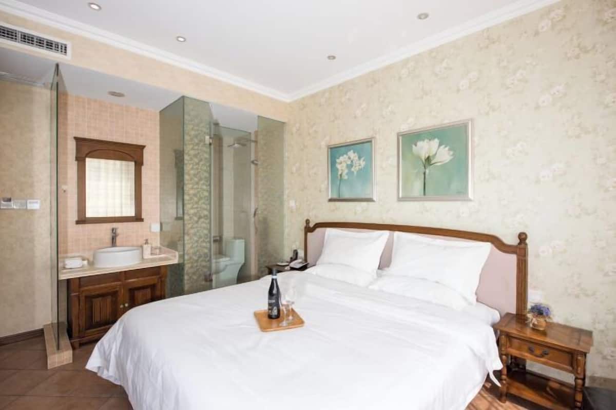 Ein Hotelzimmer mit einem großen Bett, einem hölzernen Nachttisch, einem Badezimmer mit Waschbecken und Spiegel sowie blumigem Wandpapier.