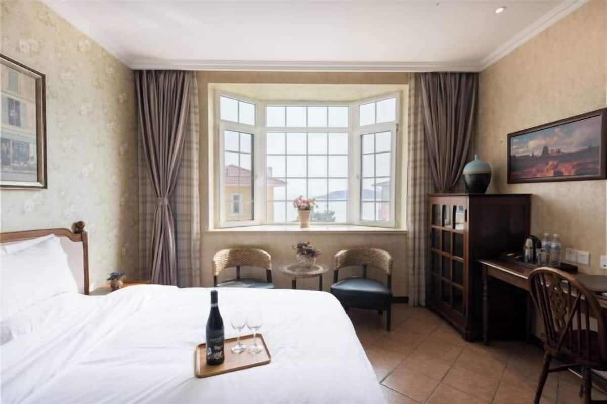 Ein Hotelzimmer mit einem großen Bett, einem Schreibtisch, zwei Stühlen, einem Fenster mit Vorhängen und einem Gemälde an der Wand.