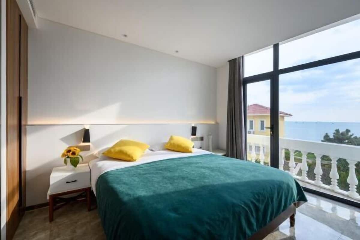 Ein Hotelzimmer mit einem großen Bett, zwei Kissen, einem Nachttisch mit Lampe und Meerblick durch große Fenster.