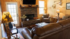A cozy log cabin interior with a stone fireplace, leather sofa, and a mounted television.