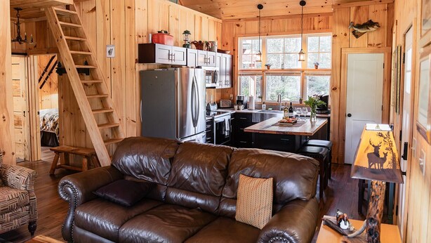 A cozy cabin interior with a leather sofa, wooden walls, a kitchen with a refrigerator and cabinets, a dining table, and a window with a view of the outdoors.