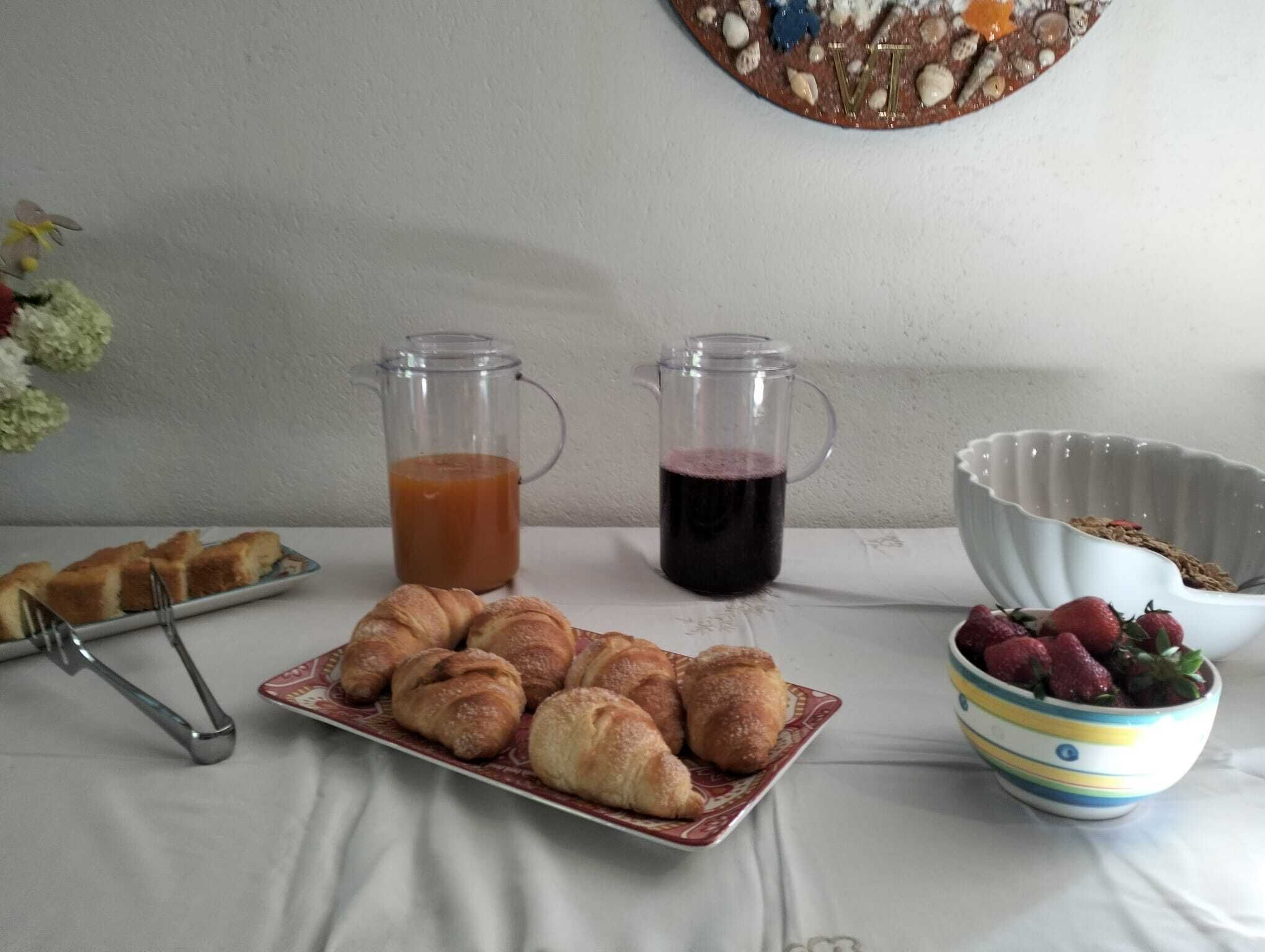 A breakfast table with croissants, two pitchers of beverages, a bowl of strawberries, and a decorative wall piece.