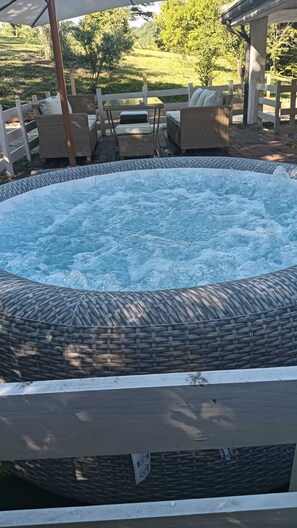 Een hottub met rieten rand, houten loungestoelen en een overdekt zitgedeelte met uitzicht op een aangelegde tuin.