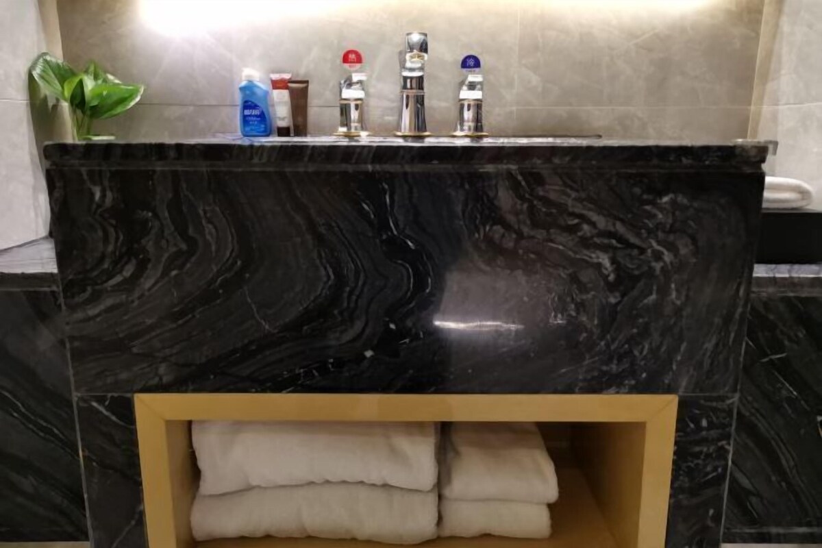 A bathroom with a black marble sink, three faucets, a plant, and a shelf with folded towels.