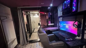 Ein Zimmer mit einer Spiele-Station, inklusive Monitor, Tastatur und Kopfhörer, einer Couch und einer Tür zu einem weiteren Raum.