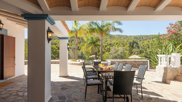Un patio al aire libre con piso de piedra, una mesa dispuesta para comer y vistas de un paisaje exuberante con árboles y colinas.