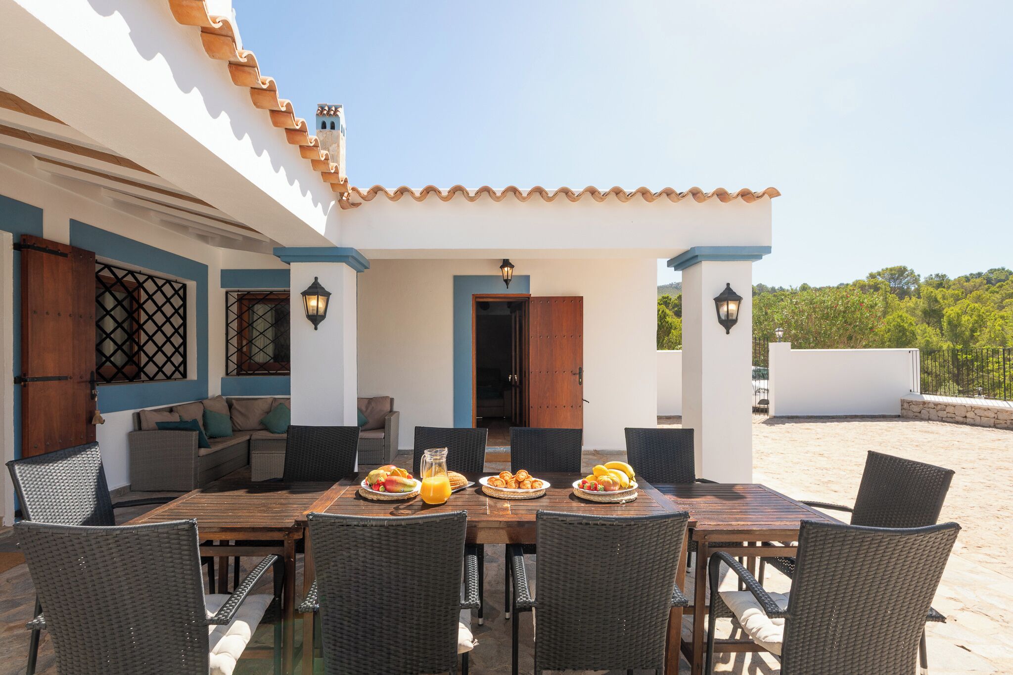 An outdoor dining area with a wooden table and wicker chairs, surrounded by a white building with blue accents and a tiled roof.