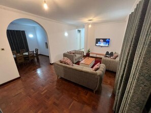 A living room with a sofa, a coffee table, and a television. There is a dining area with chairs and a table in the background. - El Tesa Court Gardens Milimani (Kisumu)