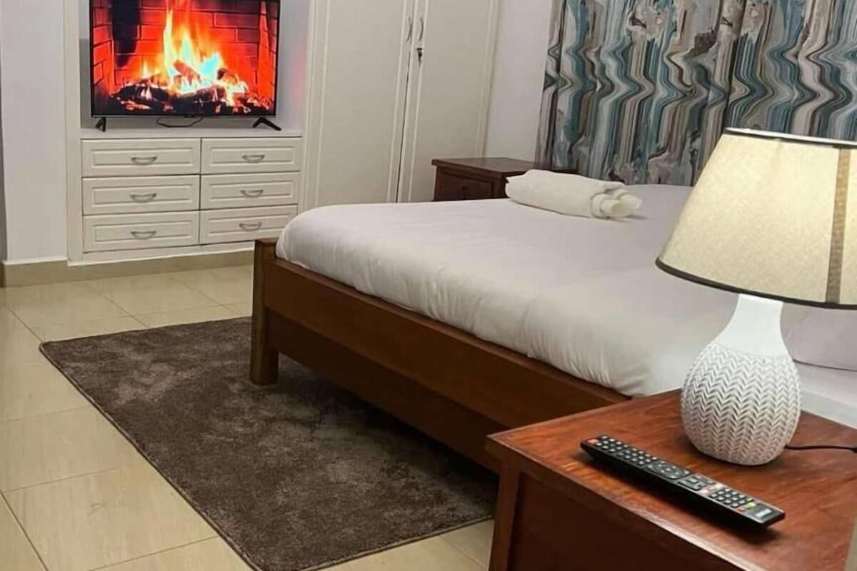 Ein Hotelzimmer mit einem Bett, einem Nachttisch mit Lampe, einem Fernseher, der einen Kamin zeigt, einem kleinen Nachttisch und einem gemusterten Vorhang.