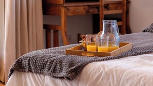Ein ordentlich bezogenes Bett mit einem Tablett Orangensaft und einer Saftkanne, ein Holztisch mit Stuhl und ein Fenster mit Vorhängen.