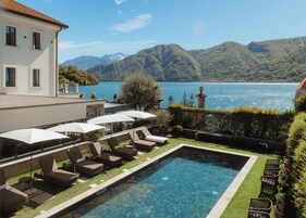 Zona de piscina con sillas de descanso y sombrillas, vista despejada de un lago y montañas al fondo.