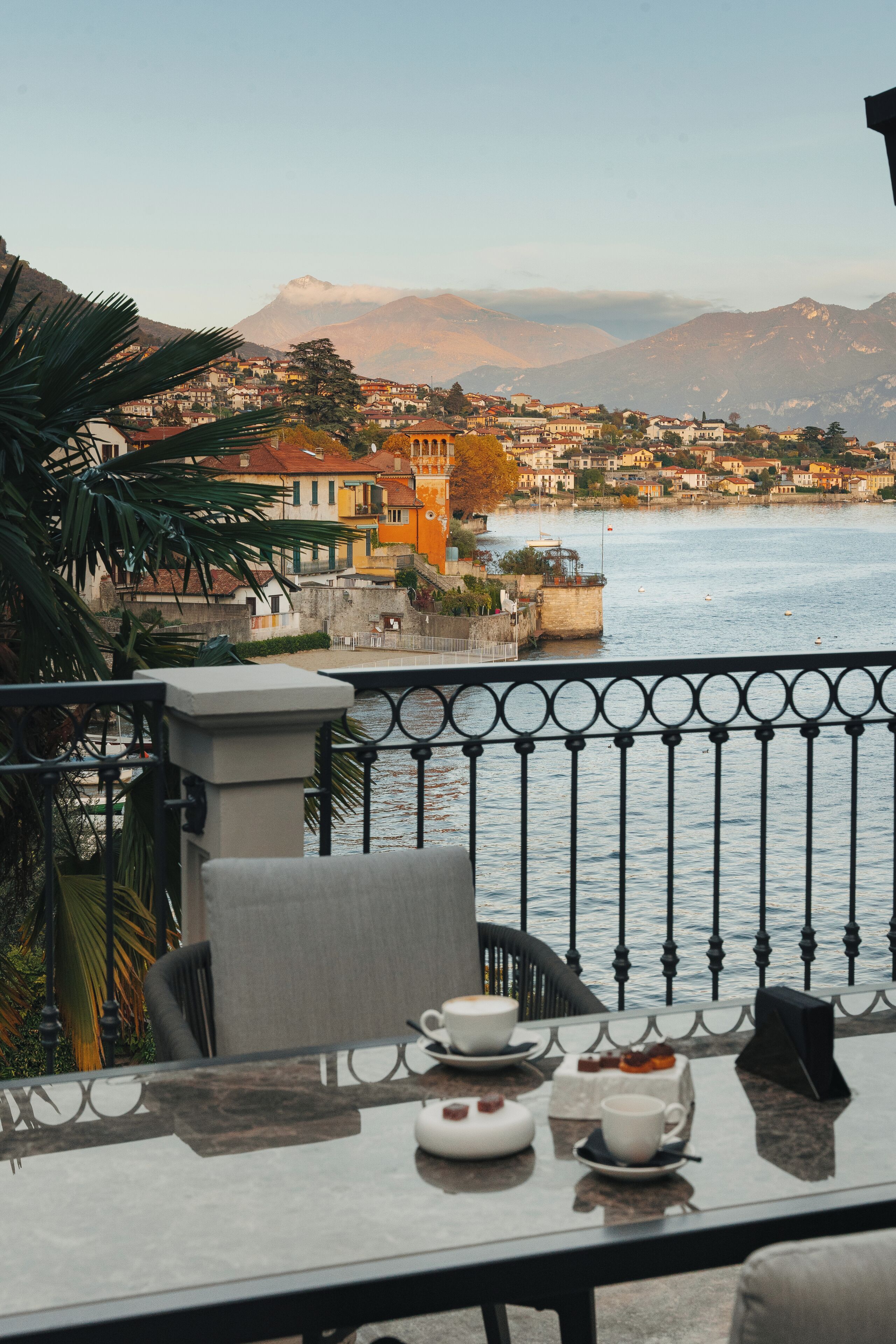 Un balcone con un tavolo apparecchiato per il tè, affacciato su un fiume con edifici e montagne sullo sfondo.