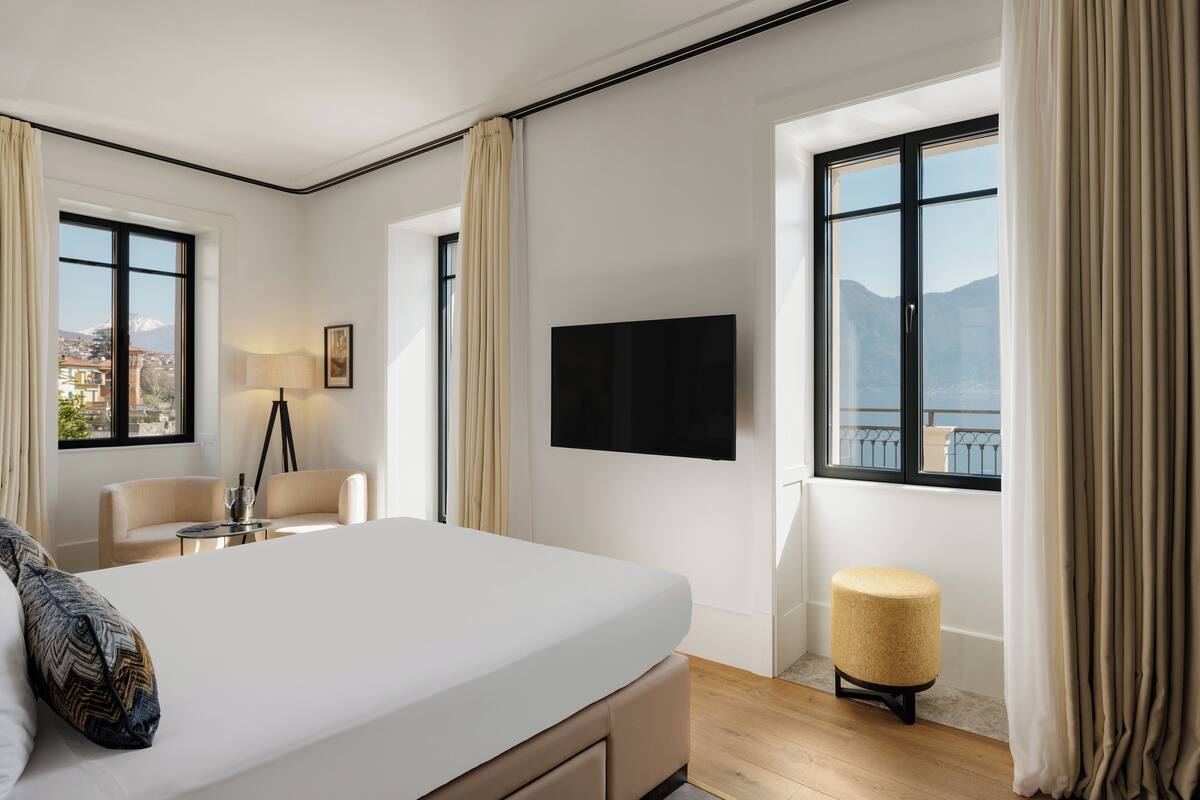 A modern hotel room with a large bed, a flat-screen TV, a small table with a lamp, and a view of the mountains through the windows.