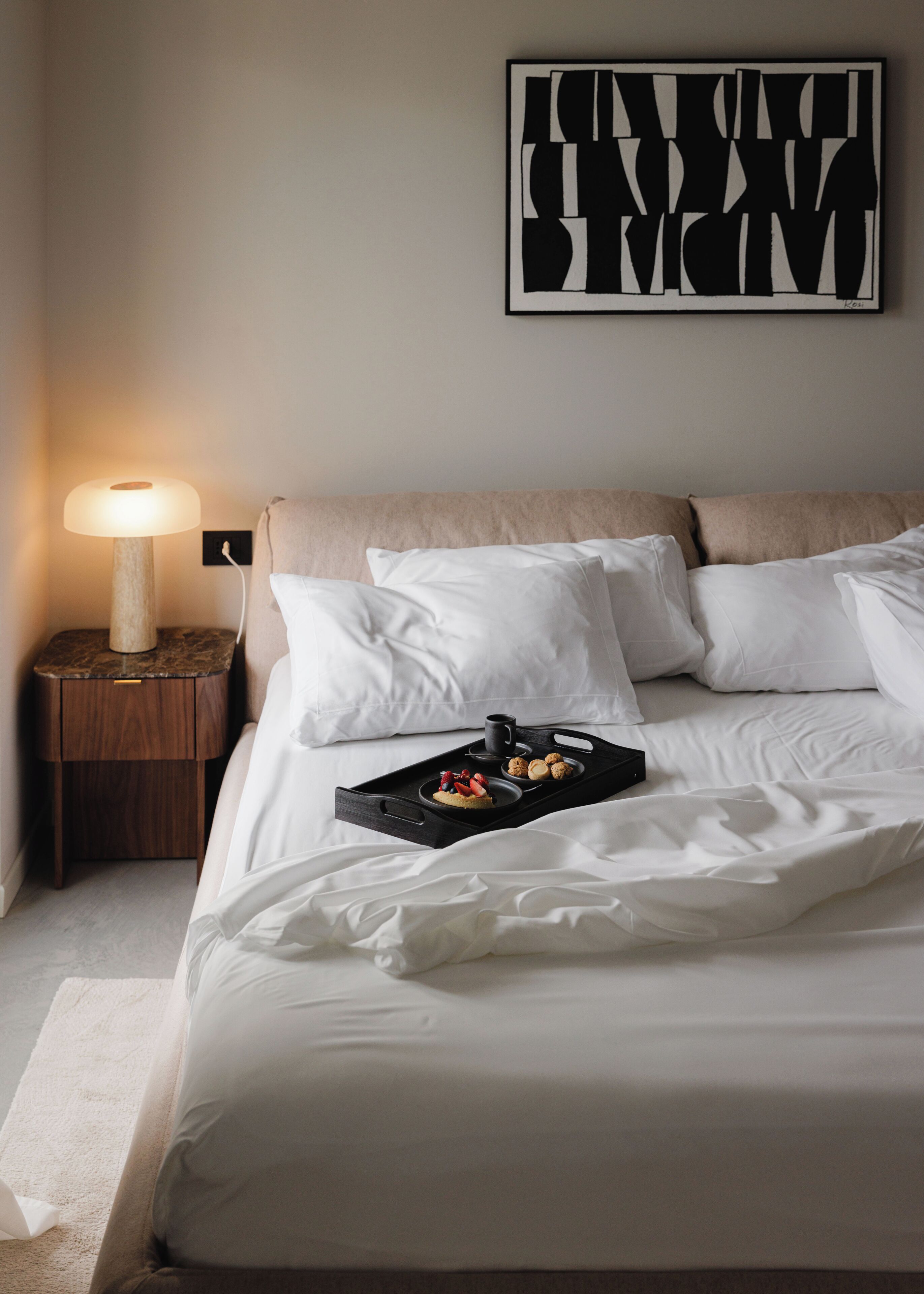 Una cama bien hecha con sábanas blancas, una mesita de noche con una lámpara y una bandeja con pasteles y una taza.