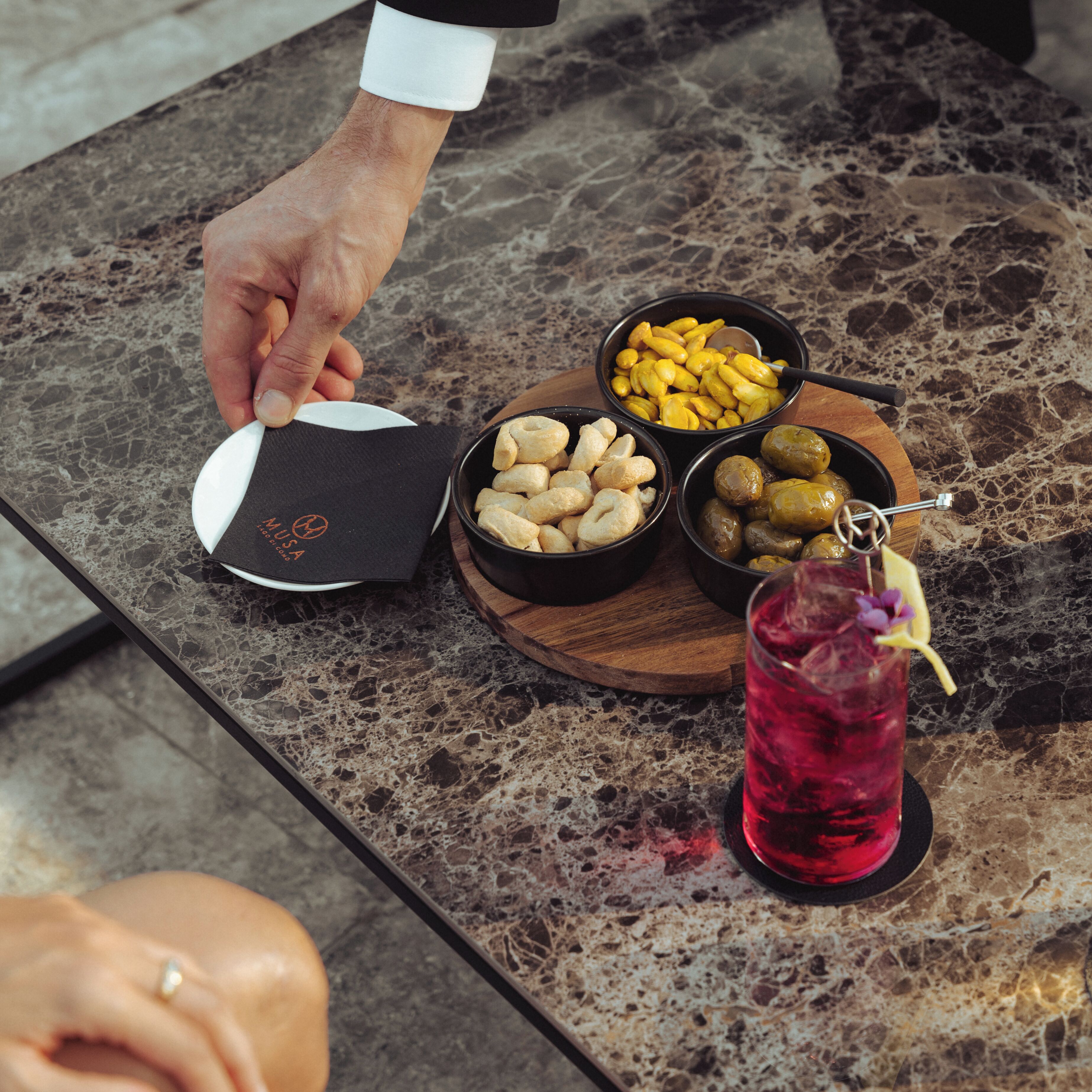 A marble table with a plate of nuts, a bowl of olives, and a glass of red drink.