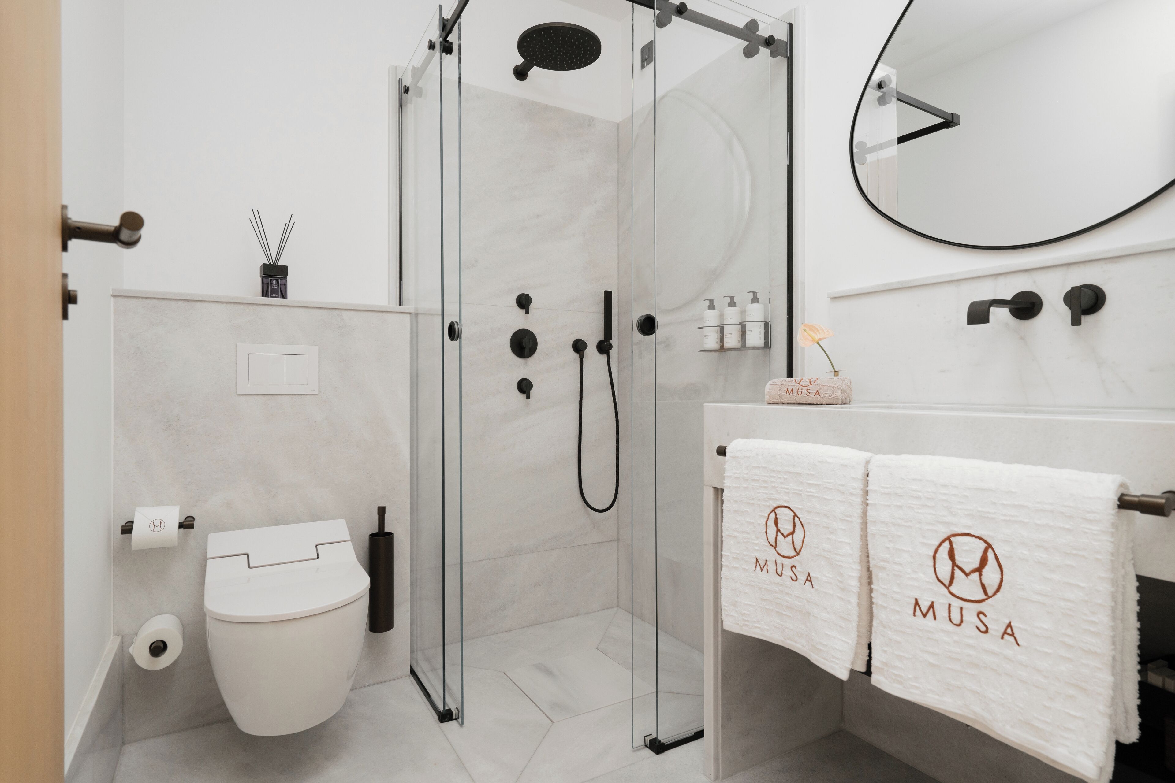Ein modernes Badezimmer mit einer gläsernen Dusche, einer Toilette, einem Waschbecken mit Spiegel und Handtüchern mit einem Logo.