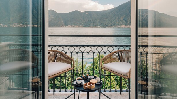 Un balcón con sillas de mimbre, una pequeña mesa redonda con una bandeja de objetos y vistas a un lago con montañas al fondo.