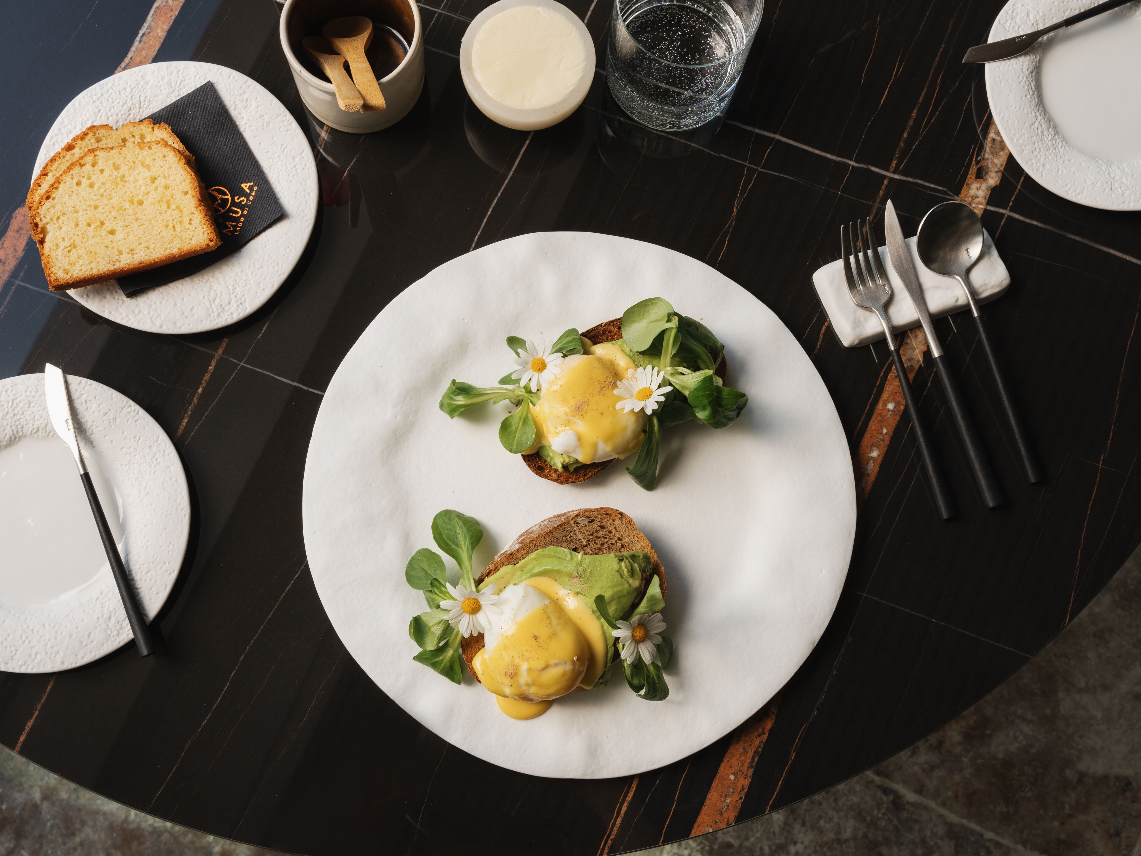 A breakfast setting with two eggs benedict sandwiches on white plates, accompanied by bread, butter, and a glass of water.