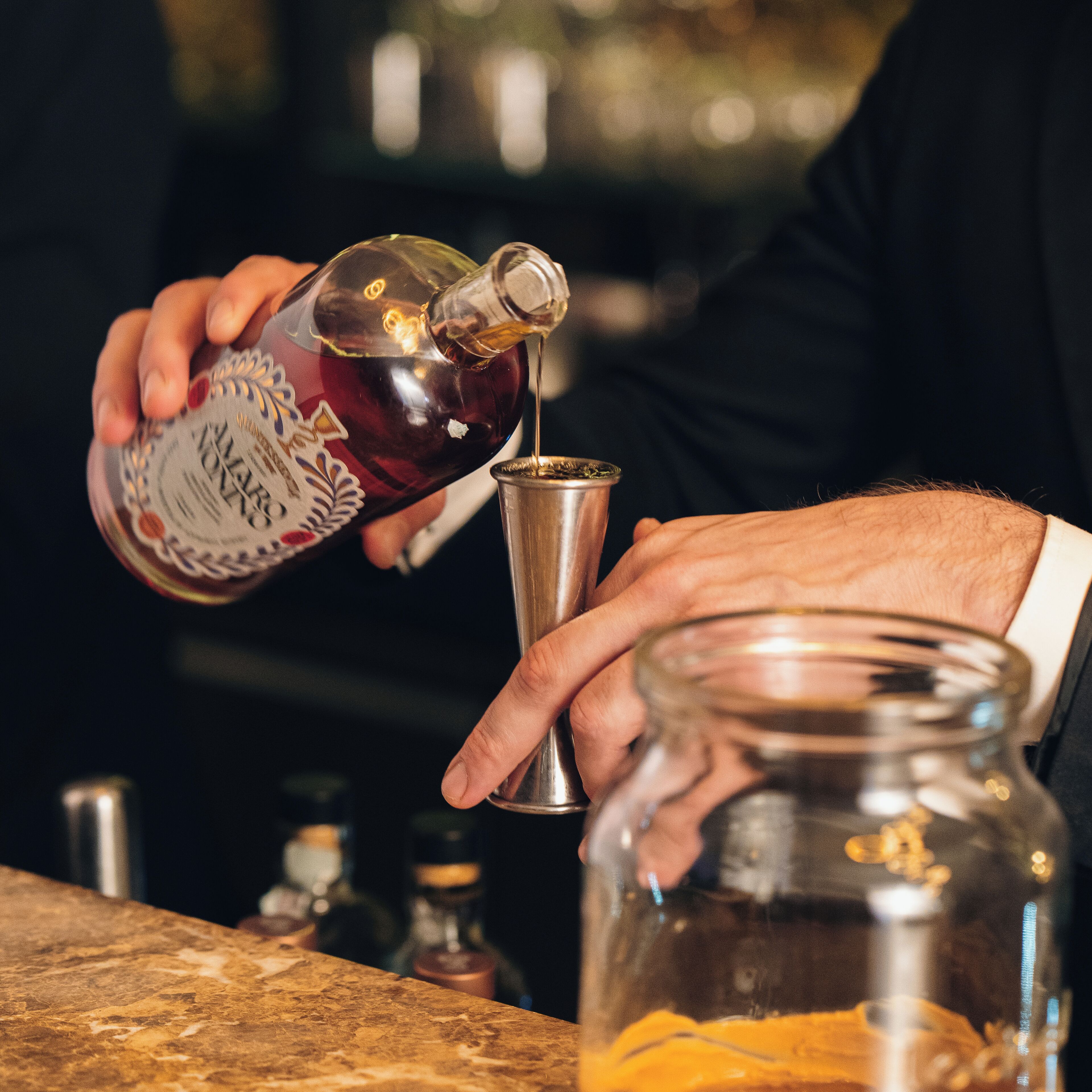 A person pouring a drink from a bottle into a metal shaker.