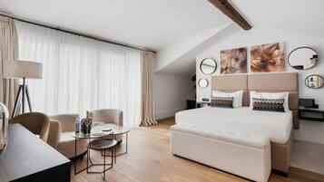 A modern hotel room with a large bed, a desk, and a chair. There are curtains, a lamp, and artwork on the wall.