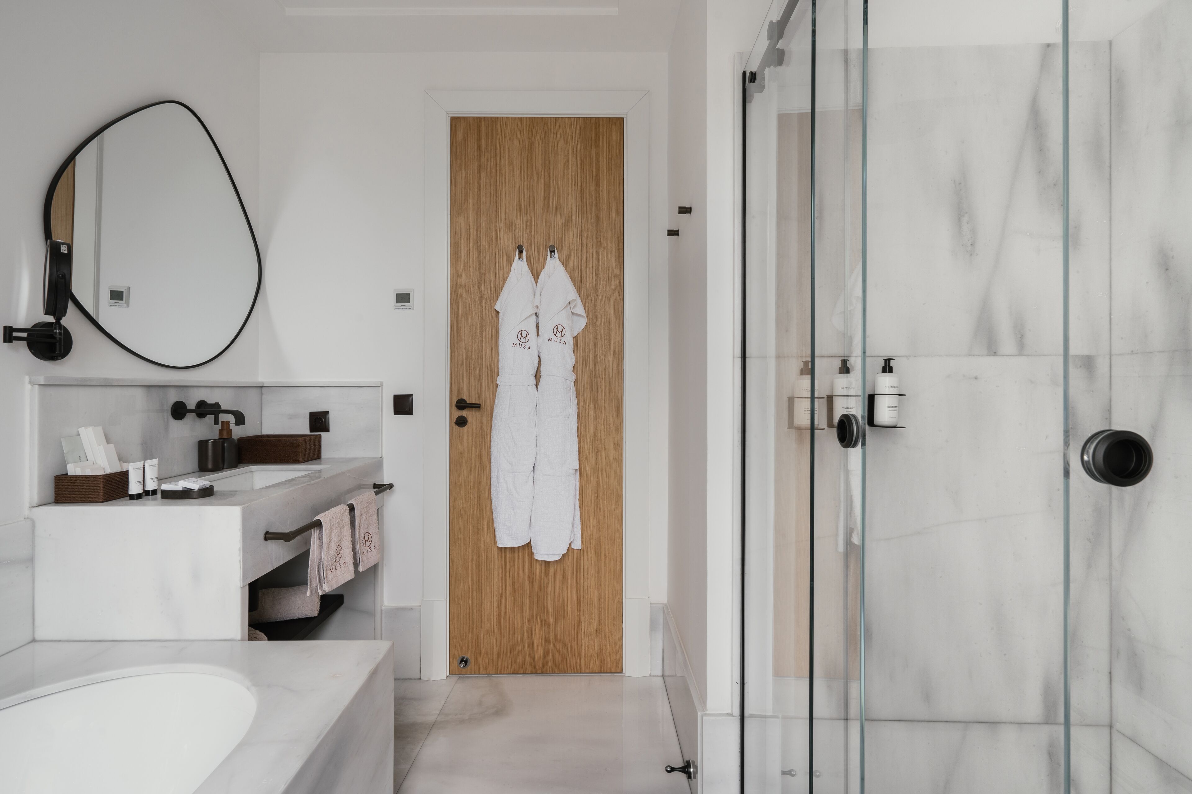 Ein modernes Badezimmer mit einem großen Spiegel, einer Holztür, einer Glasdusche und einem weißen Waschbecken.