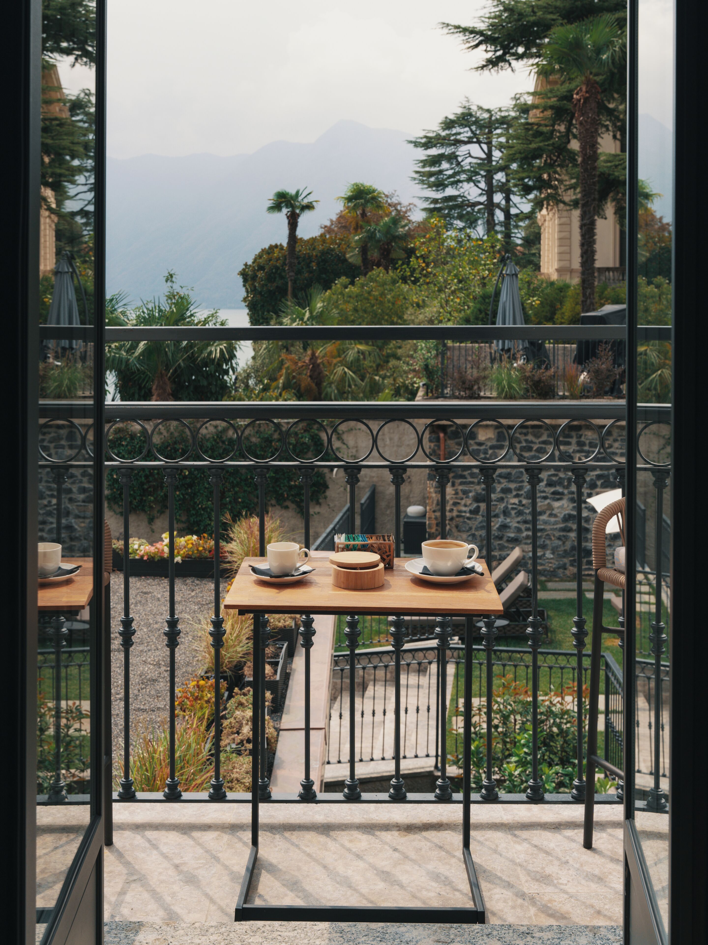 Un balcon avec une table dressée pour deux personnes, donnant sur un jardin où poussent différentes espèces de plantes et offrant une vue lointaine sur une montagne.