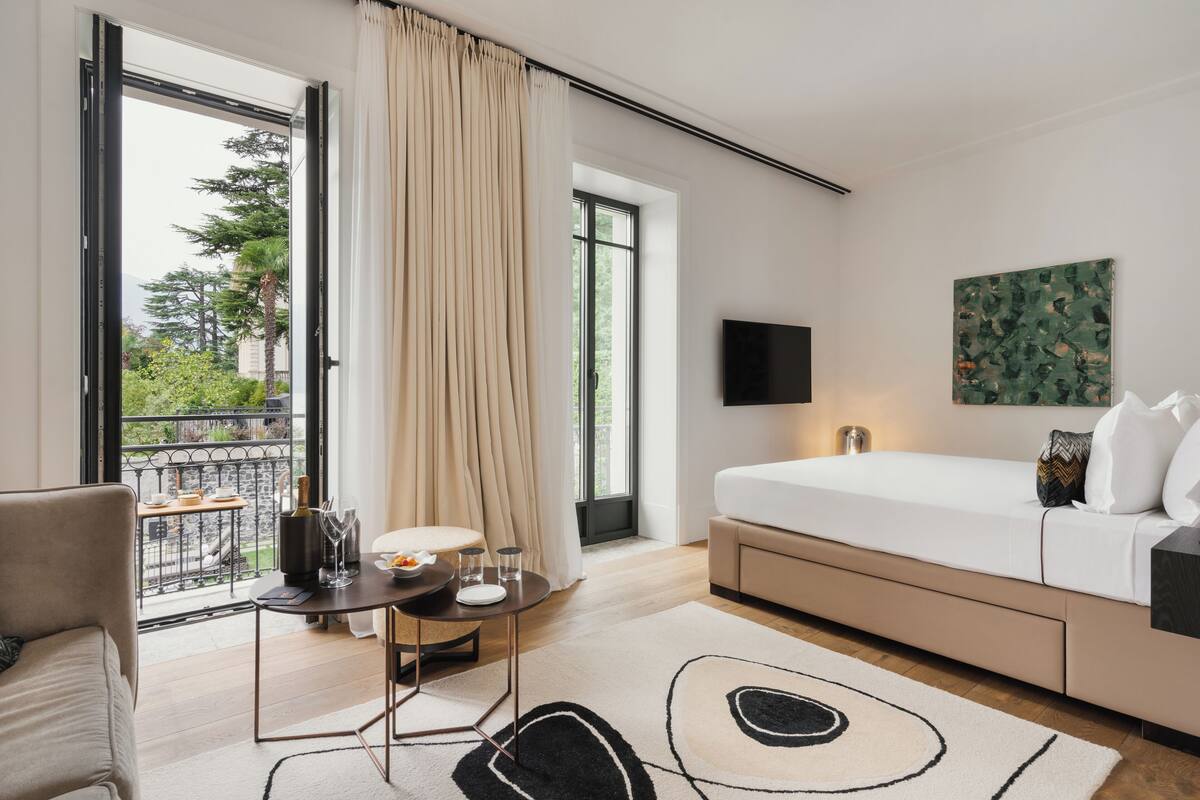 A modern hotel room with a bed, a TV, a balcony with a table and chairs, and a large window with curtains.