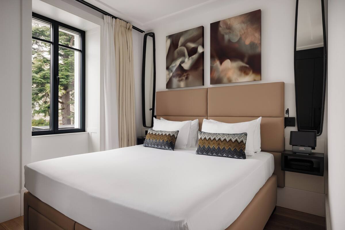 A modern hotel room with a large bed, a window with curtains, and abstract wall art.