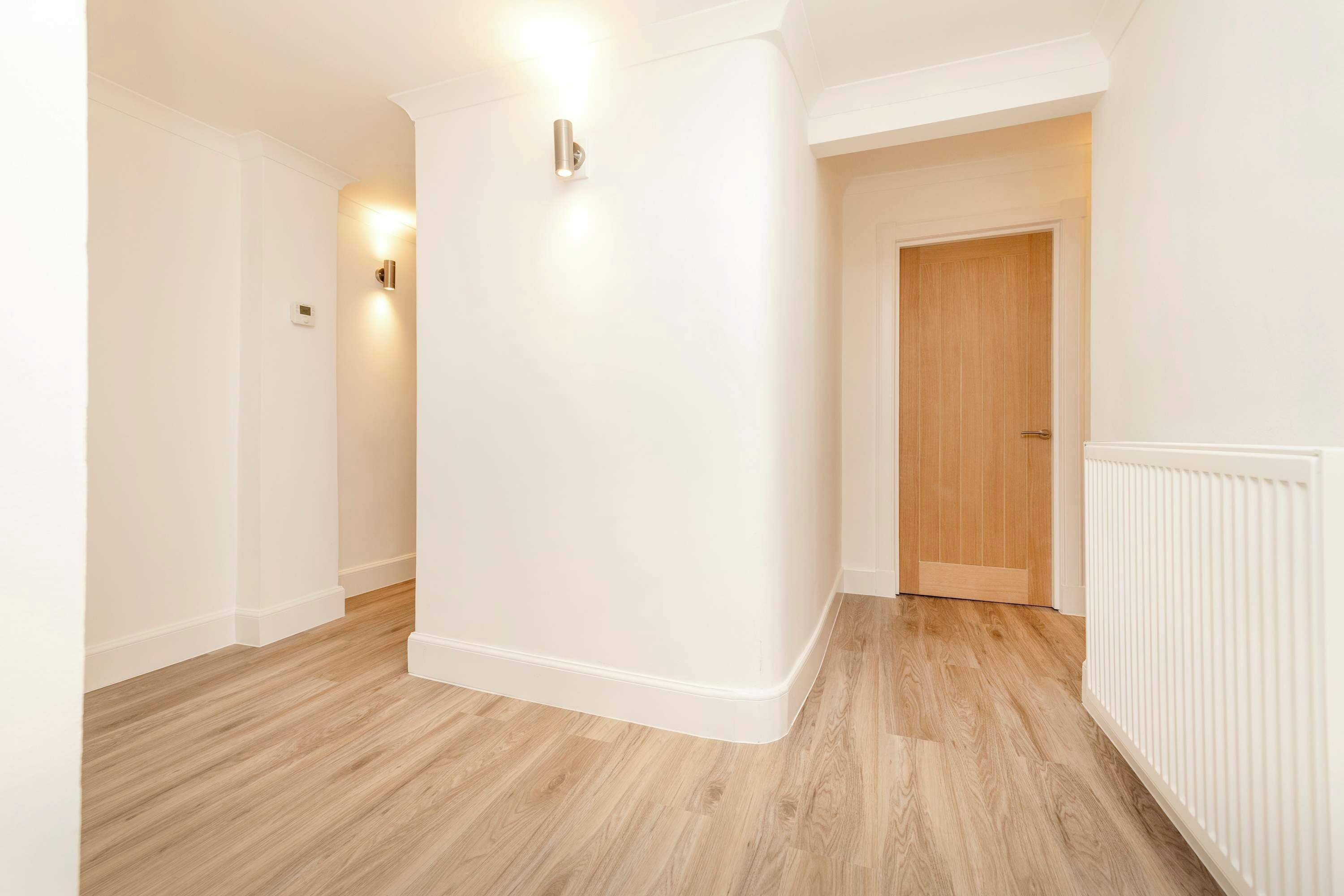 A hallway with white walls and wooden flooring, leading to a door.