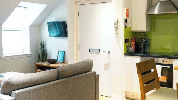 A modern living room with a grey sofa, a wooden dining table with a chair, a kitchen area with a green microwave, and a white door.