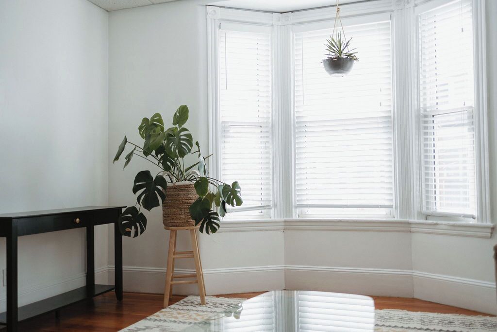 A corner room with a large window, a black table, a wooden stool, and a potted plant.