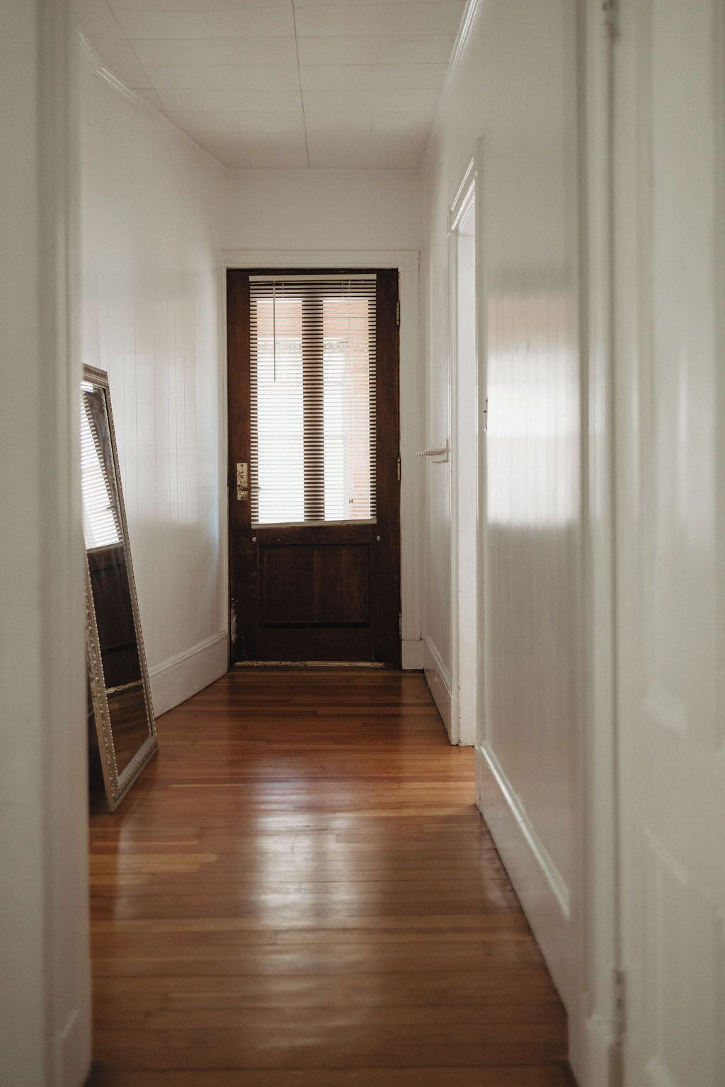 A hallway with wooden flooring, white walls, and a door with blinds.