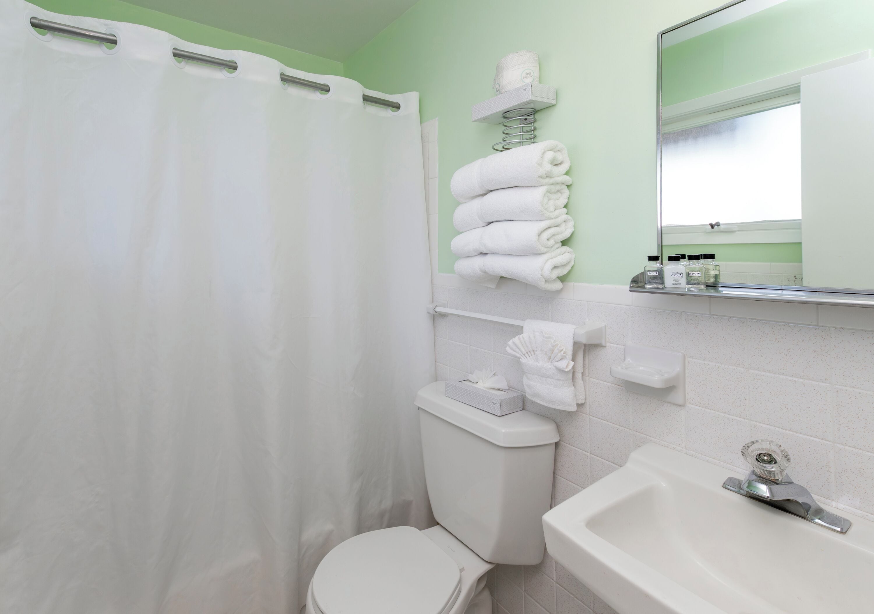 Un baño con cortina de ducha blanca, inodoro, lavamanos con espejo y estante con toallas dobladas.