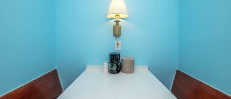 A room with a white countertop, a coffee maker, a lamp, and a trash bin against a blue wall.