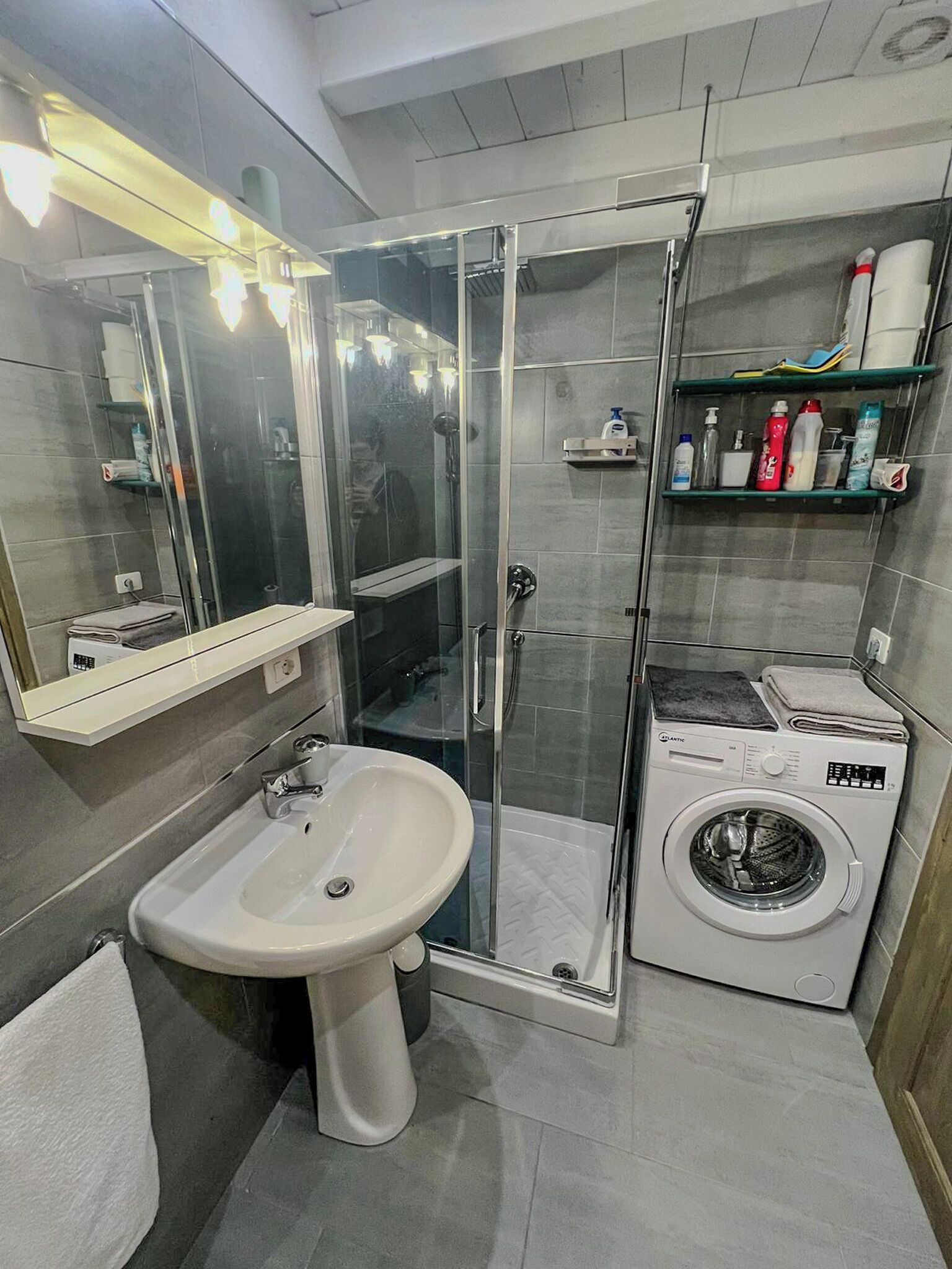 A modern bathroom with a glass-enclosed shower, a sink, a washing machine, and shelves with toiletries.