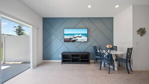 Ein modernes Wohnzimmer mit einem großen Flachbildfernseher an einer blauen Wand, einer dunklen Unterhaltungseinheit darunter und einem Essbereich mit blauen Stühlen und einem weißen Tisch.