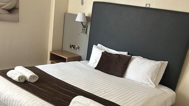 Un lit bien fait, avec une tête de lit sombre, du linge de lit blanc et deux serviettes enroulées posées sur le lit.